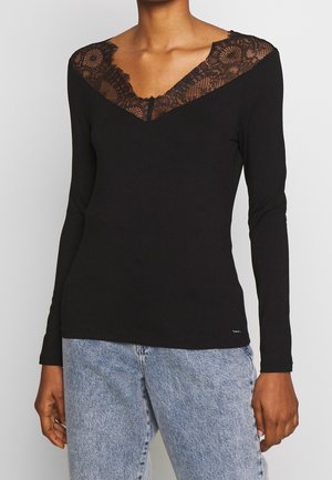 Kobieta w czarnej bluzce z długim rękawem, z koronkowym detalem dekoltu w serek, połączona z jasnoniebieskimi jeansami na jednolitym tle.
