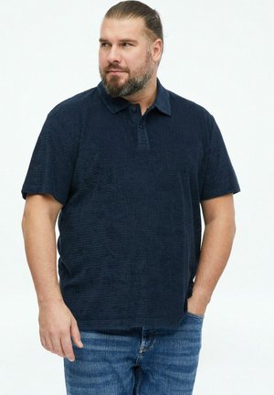 Man met baard en naar achter gebonden haar, gekleed in een marineblauw poloshirt met korte mouwen en een blauwe spijkerbroek, staand met één hand in de zak.