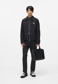 Czarna bomberka z kolnierzem wykończonym ćwiekami, zdobiona logo. Slim-fit czarne spodnie i wypolerowane czarne i białe buty. Nosi czarną, strukturalną torbę.