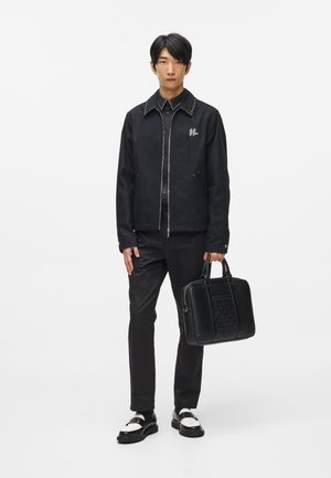 Sort bomberjakke med naglede krave, detaljeret med logo. Slim-fit sorte bukser og polerede sort-hvide sko. Bærer en sort struktureret taske.