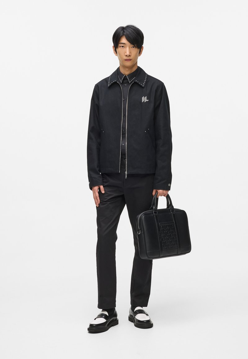 Czarna bomberka z kolnierzem wykończonym ćwiekami, zdobiona logo. Slim-fit czarne spodnie i wypolerowane czarne i białe buty. Nosi czarną, strukturalną torbę.