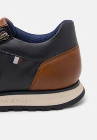 Črne in rjave usnjene čevlje z teksturiranim rjavim petnim delom, modrim notranjim oblačilom in belim podplatom. Imajo majhno oznako z zastavo blizu vezalk.