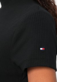 Melns ribots īss piedurkņu tops ar augstu apkakli. Kreisajā piedurknē neliels izšūts logotips sarkanā, baltā un zilā krāsā.