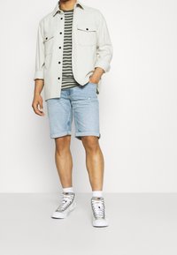 Světle šedá košile s límcem přes pruhované zelené a bílé tričko, doplněné o ohrnuté džíny šortky. Na sobě má šedé a bílé vysoké tenisky.