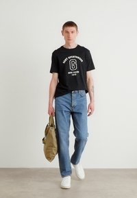 Sort T-shirt med korteærmer med hvid grafisk tekst, blå denimjeans, hvide sneakers og en tan skuldertaske. Enkel, afslappet påklædning.