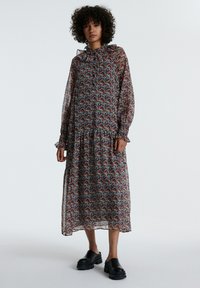 Langes, fließendes Kleid mit einem floralen Muster in Rot, Blau und Gelb auf schwarzem Hintergrund, mit langen Ärmeln und einem frillierten Saum. Schwarze Plateauschuhe.