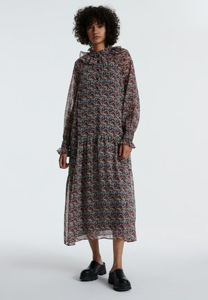 Langes, fließendes Kleid mit einem floralen Muster in Rot, Blau und Gelb auf schwarzem Hintergrund, mit langen Ärmeln und einem frillierten Saum. Schwarze Plateauschuhe.
