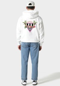 Uomo che indossa un hoodie bianco con un'auto da corsa vintage colorata e il testo "Pas de Monaco" sulla schiena, jeans blu, cappellino bianco e sneakers bianche.