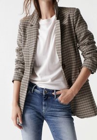 Blazer à motif pied-de-poule marron et beige avec une coupe ajustée, porté sur un t-shirt blanc et un jean bleu. Boutons distinctifs et tissu texturé.