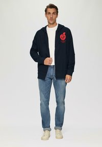 Felpa zip con cappuccio di colore blu navy, dotata di tasche frontali e grafica di una mela rossa. Indossata sopra una maglietta bianca, abbinata a jeans azzurri chiari e sneakers beige.