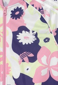 Adidas zip-up jacka med blommönster i rosa, lila och vitt, med textilstruktur och broderad logotyp på bröstet.