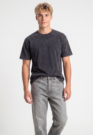 Giovane uomo con capelli biondi ricci che indossa una t-shirt scura slavata e jeans grigio chiaro, in piedi davanti a uno sfondo bianco semplice.