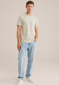 Svetlo béžové tričko s okrúhlym výstrihom vyrobené z jemnej bavlny, skombinované so svetlo modrými rovnými džínsy a bielymi teniskami, stojaci na jednoduchej pozadí.