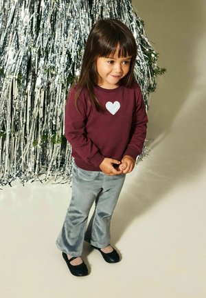 Bordowa koszula z długim rękawem z srebrnym wzorem serca, zestawiona z jasnoszarymi rozszerzanymi spodniami i czarnymi butami. W tle dekoracje w metalicznym stylu.