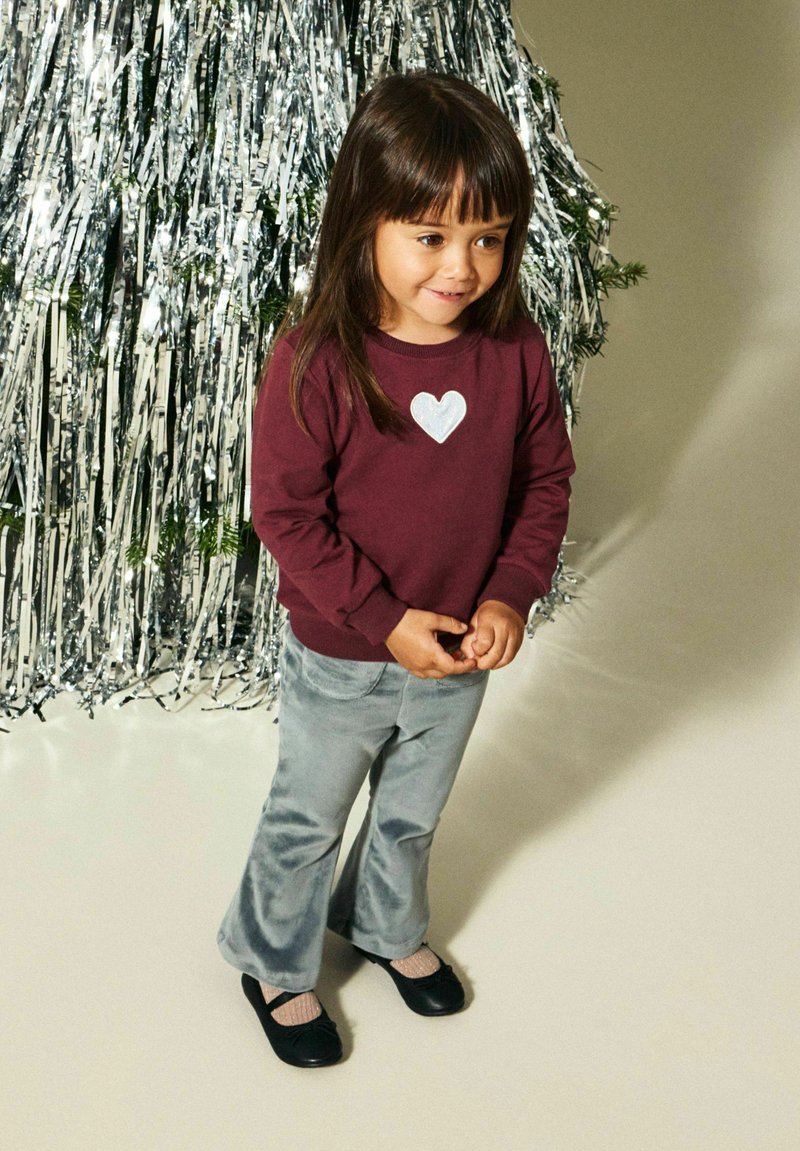 Chemise bordeaux à manches longues avec un motif de cœur argenté, associée à un pantalon évasé gris clair et des chaussures noires. Le fond présente une décoration métallique.