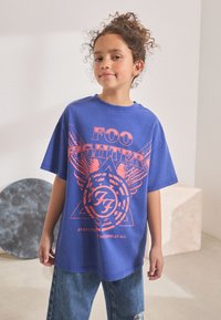 Modra prevelika majica s kratkimi rokavi z oranžnim grafičnim potiskom, ki prikazuje krila in okroglo obliko. Nosi se s strganimi jeansi iz džinsa.