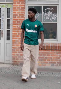 Grön Puma sporttröja med "Auchan"-logotyp, beiga vida byxor och vita sneakers. Texturerad tegelvägg i en stadsmiljö.