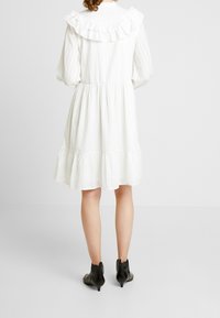 Vit bomulls klänning med volangkrage, långa ärmar och lager-på-lager nederkant. Matchad med svarta ankelskor. Texturerad tyg är synlig.