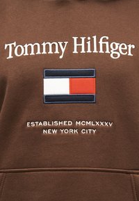 Καφέ ύφασμα με κεντημένο κείμενο "Tommy Hilfiger," το κόκκινο, λευκό και μπλε λογότυπο της σημαίας της μάρκας, και "Established MCMLXXXV New York City."