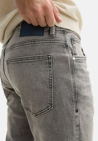 Szürke farmernadrág, amely illeszkedő fazonú, hátul zsebekkel, kiemelt öltéssel és sötét bőr címkével a derékvonalon.