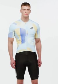 Kurzarm-Radtrikot in Hellblau mit abstrakten gelben und lavendelfarbenen Mustern, kombiniert mit eng anliegenden schwarzen Shorts mit weißen Streifen.