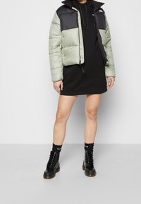 Pufferjacka i ljusgrön med en svart överdel, med dragkedja framtill och sidofickor. Bärs över en svart sweatshirtklänning och svarta stövlar.