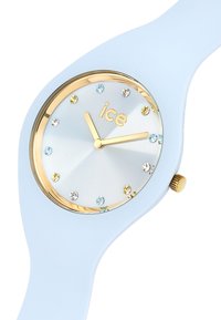 Montre en silicone bleu clair avec un cadran doré, un cadran argenté et des index en cristal. Présente un logo doré et une couronne sur le côté.