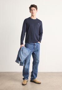 Navy long-sleeve shirt with crew neck, paired with light blue denim jeans. Model holding a denim jacket. Beige shoes. Neutral background.