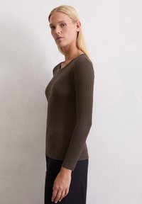 Bruine geribbelde langemouwen top met een ronde hals, slank ontwerp, zachte textuur en een rechte zoom, gedragen met zwarte broek, poseren tegen een lichte muur.