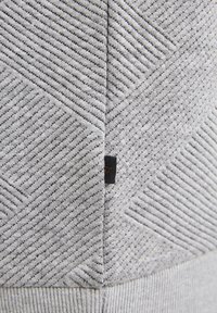 Grijze gebreide muts met diagonale textuurpatronen, voorzien van een klein zwart label aan de naad. Zacht materiaal met een comfortabele pasvorm.