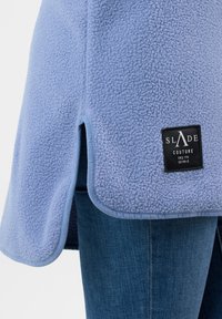 Wrap in felpa azzurra chiara con fessure laterali, caratterizzato da un'etichetta nera con la scritta "SLADE COUTURE." Testura morbida e bordi arrotondati alle aperture.