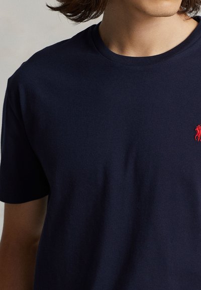 Person iført en marineblå t-shirt med en lille rød broderet logo på venstre bryst, der viser nakke, skulder og del af armen.