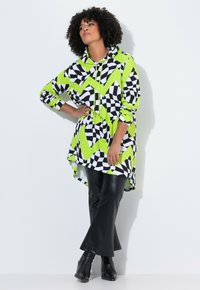 Donna in pantaloni in ecopelle neri e stivali neri, che indossa un hoodie largo con motivo a zigzag in verde neon, nero e bianco, in piedi con una mano sulla vita.