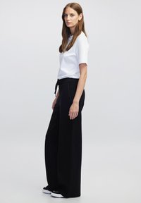 Zwarte wijde broek met een trekkoord in de taille, gecombineerd met een aansluitend wit t-shirt. Kenmerkt zich door eenvoudige, strakke lijnen en een ontspannen pasvorm.