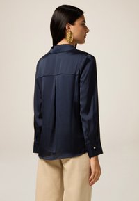 Marineblaue Satinhose mit langen Ärmeln, Kragen, Rückendetail mit Falte und geknöpften Manschetten, kombiniert mit beigen Hosen.