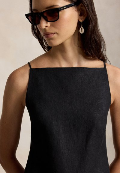 Γυναίκα που φοράει μαύρο τοπ χωρίς μανίκια με τετράγωνη λαιμόκοψη, σκούρα γυαλιά ηλίου και σκουλαρίκι σε σχήμα κοχυλιού, με μακριά σκούρα μαλλιά.