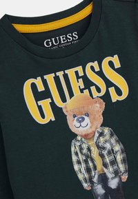 Tamsiai žalia medvilninė marškinėliai su lokio grafika geltoname kepurėje, su languotu švarko raštu ir „GUESS“ tekstu ryškiomis geltonomis raidėmis.