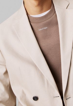 Personne portant un blazer beige par-dessus un pull Calvin Klein marron avec un lettrage blanc près de l'encolure.