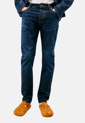 Jeans in denim blu scuro slim-fit con dettagli di cucitura arancioni, abbinati a scarpe slip-on in suede marroni con accentuazione a fibbia.