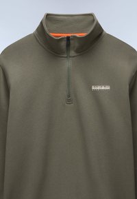 Olivengrøn kvart-zip trøje lavet af blødt stof, med en kontrasterende orange inderside ved halsen og broderet logo på brystet.