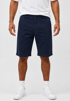 Man draagt marineblauwe knielange shorts, witte sportsokken, witte sneakers en een ingestoken wit T-shirt, staand tegen een effen achtergrond.