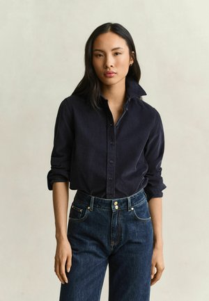 Jeune femme aux longs cheveux foncés porte une chemise sombre à boutons avec manches retroussées et un jean bleu foncé taille haute sur un fond uni.