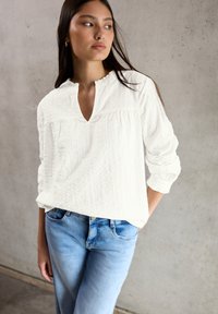 Blusa bianca, di tessuto testurizzato, con un colletto piccolo e maniche ampie, abbinata a jeans in denim blu chiaro. Design semplice e casual.