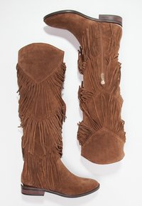 Las botas de ante marrón de caña alta cuentan con detalle de flecos a lo largo de los lados y un cierre de cremallera en el interior. Diseñadas con suela plana y punta redondeada.