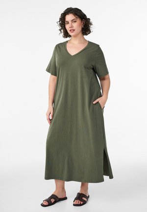 Kurdsete juustega naine, kes kannab oliivrohelise V-kaelusega lühikeste varrukatega maksikleit ja musti libisevaid sandaale, seisab ning hoiab ühte kätt taskus.