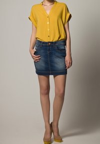 Żółta bluzka z krótkim rękawem, dekoltem w kształcie litery V i zapięciem na guziki, zestawiona z ciemną ołówkową spódnicą z dżinsu. Modelka ma na sobie beżowe szpilki.