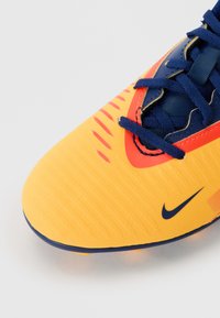 Chaussure de football Nike avec une surface jaune vif, accents bleu marine distinctifs, tige texturée avec des lignes horizontales et des détails rouges.