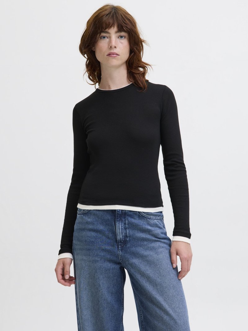 Mladá žena s hnedými vlasmi po plecia, ktorá má na sebe čierny dlhý rukáv vrchného odevu preložený cez bielu košeľu a modré džínsy, stojí pred bielym pozadím.