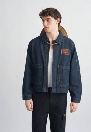 Fiatal férfi sötét farmerdzsekiben, melyen nagyelső zsebek és egy "Dickies" felvarró található, fekete nadrággal, egyszínű háttér előtt.