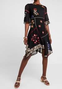 Musta mekko, jossa on kukka-ompeleita, röyhelöinen helma, lyhyet hihat ja korkea-matala leikkaus. Kevyt kangas, jossa yksityiskohtaiset kuviot.